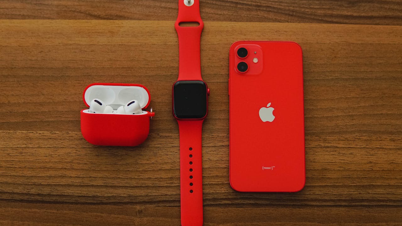 Top view flat lay of red apple watch, iPhone, and AirPods on wooden table.